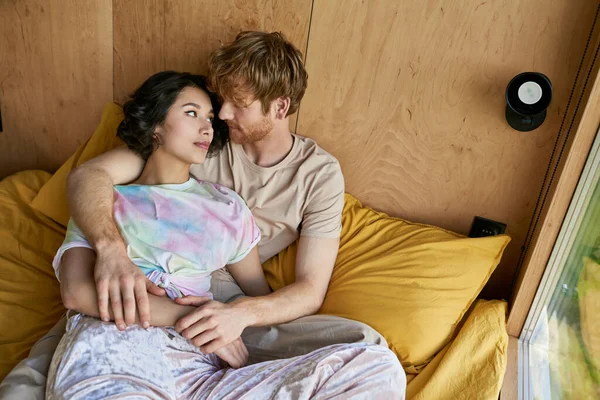 Vue de dessus, rousse homme embrassant jolie femme asiatique et couché sur le lit près de la fenêtre dans la maison de campagne — Photo de stock
