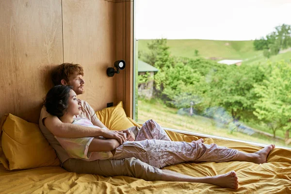 Мультикультурна пара надувається і насолоджується видом з вікна з зеленими деревами, заміський будинок — стокове фото