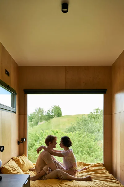 Escapadela romântica, casal multicultural olhando um para o outro enquanto sentado na cama na casa de campo — Fotografia de Stock