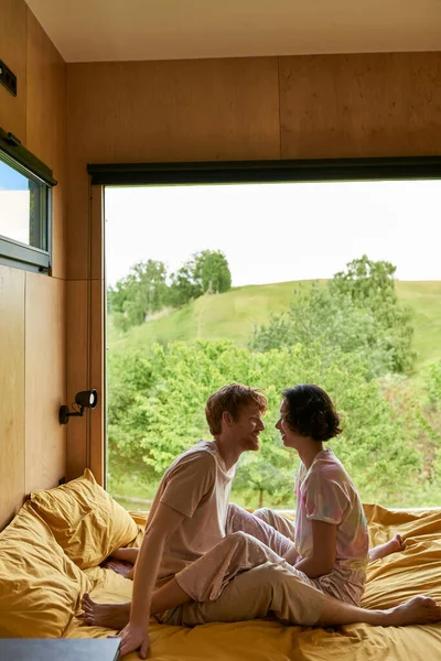 Feliz casal diverso olhando um para o outro e sentado na cama na casa de campo com vista para a floresta — Fotografia de Stock