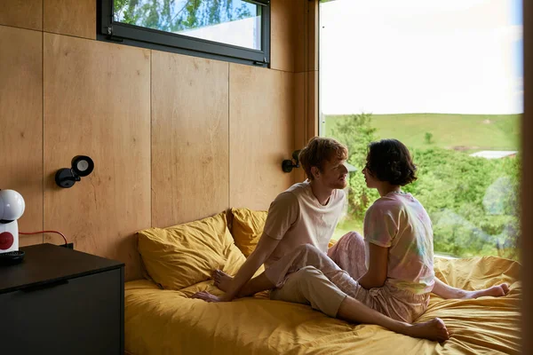 Мультиэтническая пара смотрит друг на друга и сидит на кровати в загородном доме с видом на лес — стоковое фото