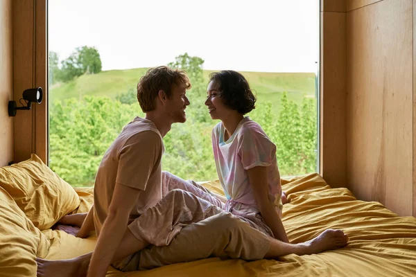 Радісна багатоетнічна пара дивиться один на одного і сидить на ліжку в заміському будинку з видом на ліс — стокове фото