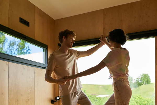 Despreocupados pareja interracial tomados de la mano y bailando juntos en casa, feliz hombre y mujer - foto de stock