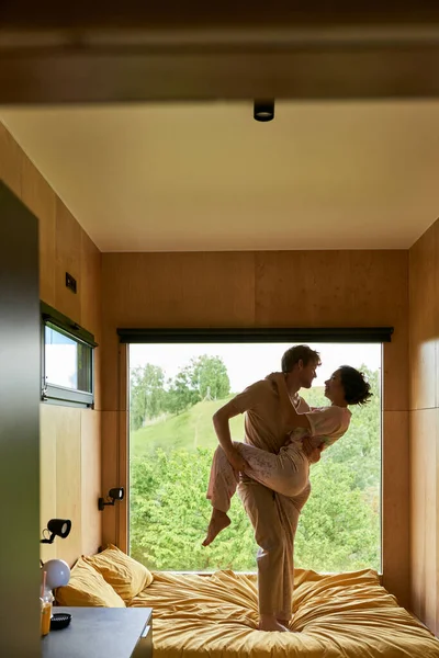 Безтурботна мультикультурна пара танцює разом на ліжку в заміському будинку, вікно з видом на ліс — стокове фото