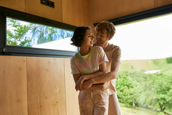 Радісний рудий чоловік обіймає азіатську дівчину біля вікна з видом на ліс в заміському будинку — стокове фото