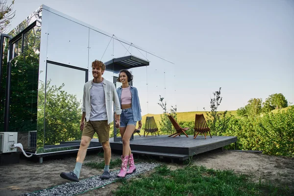 Heureux couple multiculturel marchant ensemble près de la maison de verre moderne dans la campagne, emplacement naturel — Photo de stock