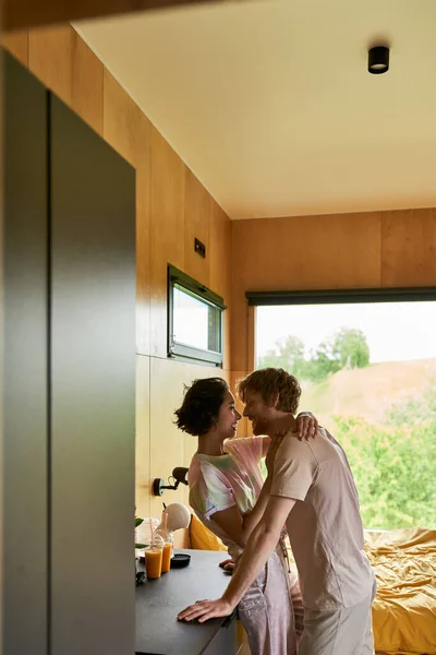 Glückliches multikulturelles Paar umarmt, während es in der Küche des Landhauses mit Blick auf den Wald steht — Stockfoto