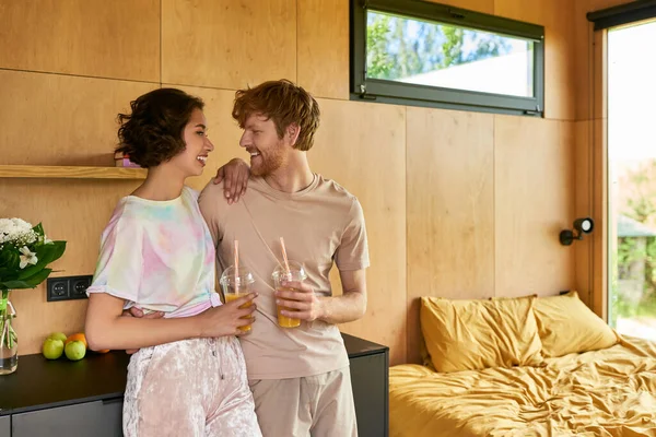 Positiva coppia diversificata in piedi in pigiama e tenendo i bicchieri con succo d'arancia al mattino — Foto stock