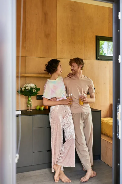 Радостная босоногая пара, стоящая в пижаме и держащая стаканы с апельсиновым соком утром — стоковое фото