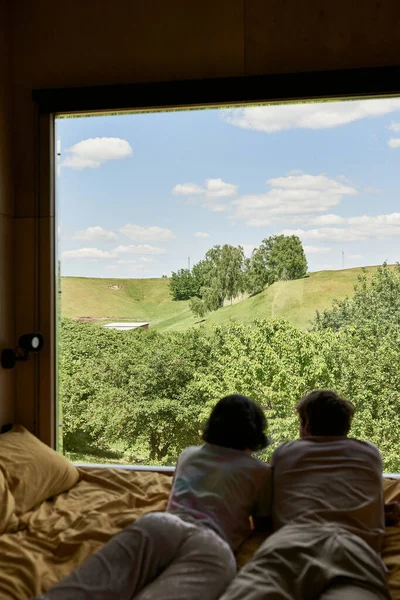 Vue panoramique, vue arrière du couple couché sur le lit et regardant les arbres verts sur la colline derrière la fenêtre — Photo de stock