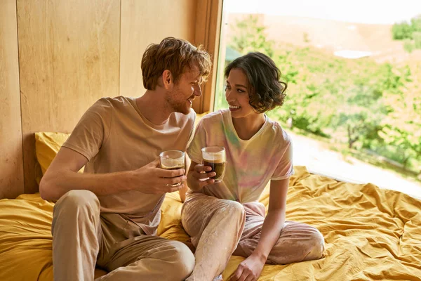 Щаслива міжрасова пара сидить разом на ліжку і тримає чашки ранкової кави, відпочинок на вихідних — стокове фото