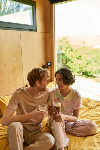 Позитивная межрасовая пара сидя на кровати и держа чашки утреннего кофе, выходные вибрации — стоковое фото