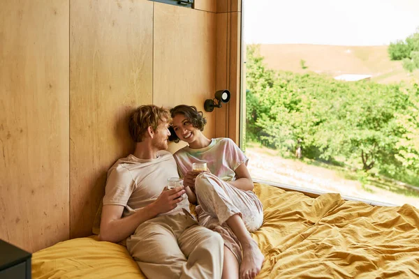 Glückliches gemischtrassiges Paar sitzt auf dem Bett und hält Tassen mit Morgenkaffee im Landhaus — Stockfoto
