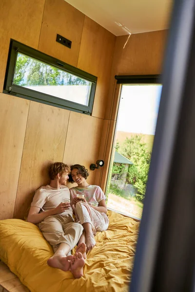 Allegra coppia interrazziale seduta sul letto e con in mano tazze di caffè del mattino in casa di campagna — Foto stock