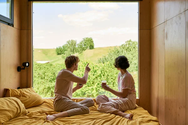 Вид ззаду людини, що вказує на зелені дерева за вікном, пара тримає чашки кави в спальні — стокове фото