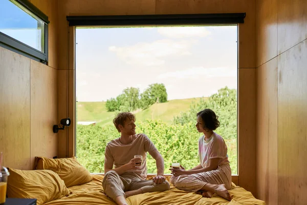 Міжрасовий пара тримає чашки кави і сидить на ліжку поруч з вікном з красивим видом — стокове фото