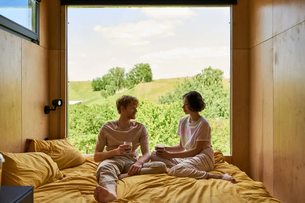 Feliz casal diverso segurando xícaras de café e sentado na cama ao lado da janela com bela vista — Fotografia de Stock