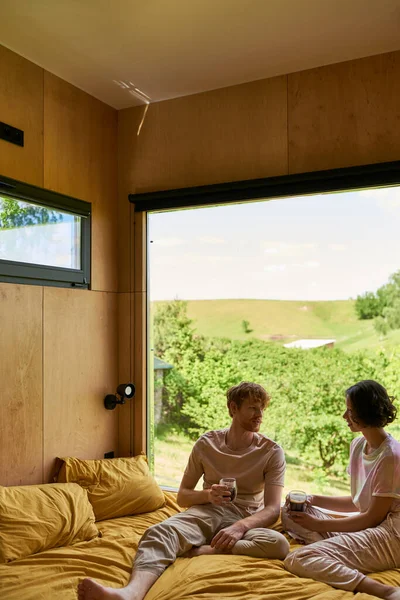Multikulturelles Paar hält Tassen Kaffee in der Hand und sitzt auf dem Bett neben dem Fenster mit schöner Aussicht — Stockfoto