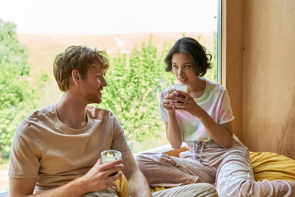 Мультикультурная пара держит чашки кофе и сидит на кровати рядом с окном с естественным видом — стоковое фото