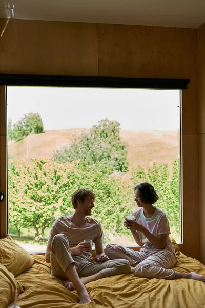 Щаслива багатоетнічна пара тримає чашки кави і сидить на ліжку біля вікна з природним видом — стокове фото