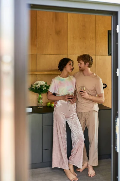 Beautiful interracial couple standing in pajamas and holding cups with morning coffee in kitchen — Stock Photo