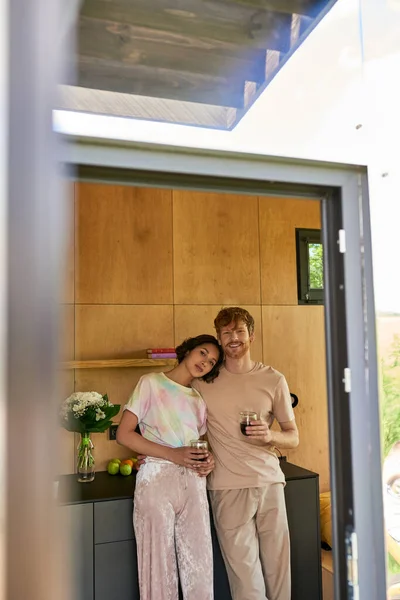 Beau couple multiethnique debout en pyjama et tenant des tasses avec café du matin dans la cuisine — Photo de stock