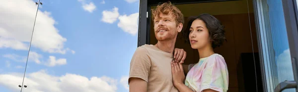 Dreamy interracial couple standing at front door of modern glass house and looking away, banner — Stock Photo