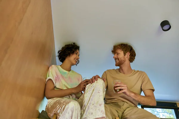 Happy asian woman and redhead man holding cups of coffee and sitting together at cozy bunk bed — Stock Photo