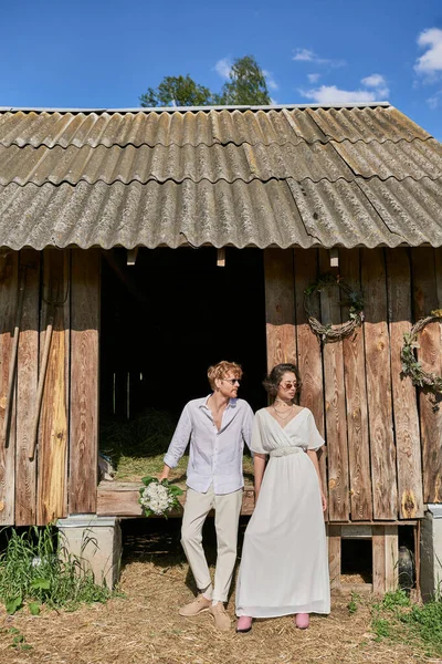 Countryside wedding concept interracial newlyweds in sunglasses and wedding gown near wooden barn — Stock Photo