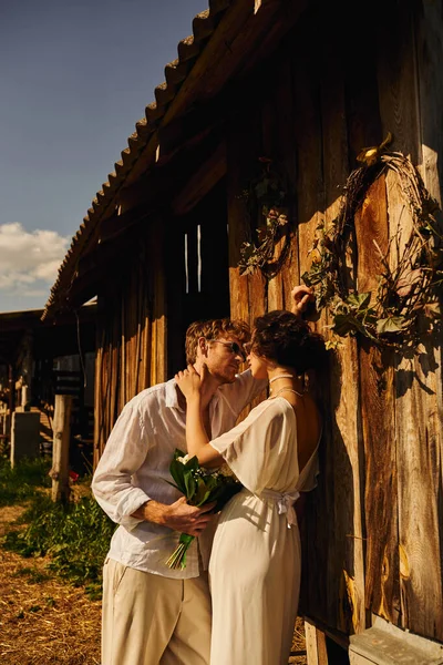 Redhead man in sunglasses holding bouquet near pretty asian woman in wedding dress near wooden barn — Stock Photo
