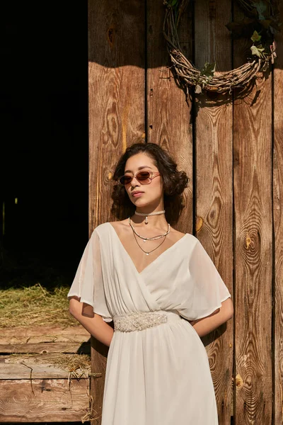 Jeune mariée asiatique en robe de mariée blanche et lunettes de soleil debout près de la grange en bois dans la campagne — Photo de stock