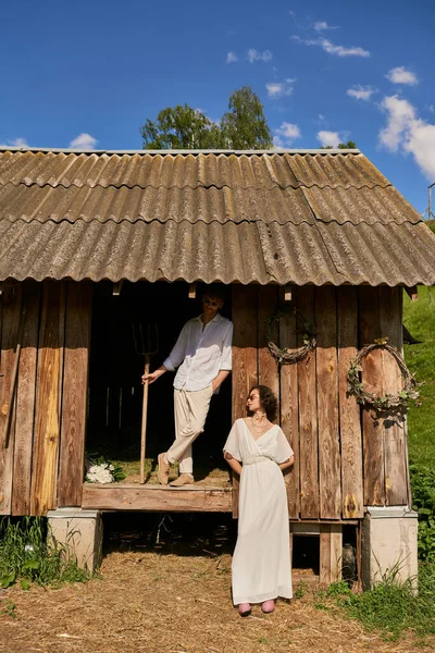 Concepto de boda rústico, interracial recién casados posando cerca de granero de madera, pareja en vestido de novia - foto de stock