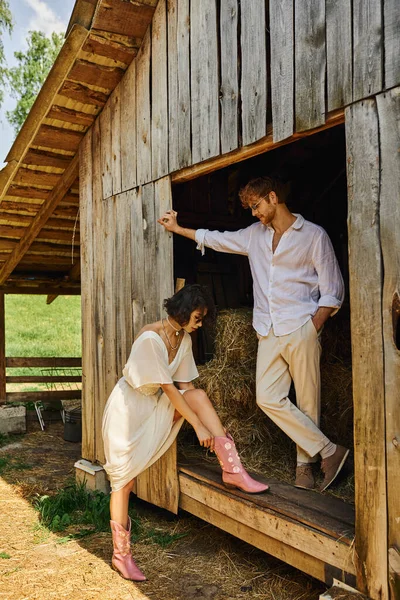 Elegante asiático novia usando rosa vaquero botas cerca pelirroja novio mientras de pie cerca de madera granero - foto de stock