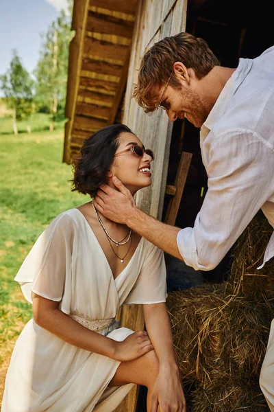 Happy redhead groom touching face of beautiful asian bride in white dress near wooden barn — Stock Photo