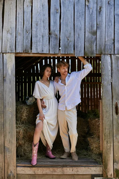 Recién casados en el campo, novia asiática en botas de vaquero y vestido blanco de pie con el novio en el granero - foto de stock