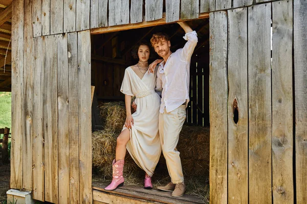 Stylish newlyweds, pretty asian bride in cowboy boots and white dress standing with groom in barn — Stock Photo