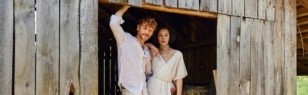 Recién casados en el campo, novia asiática en vestido blanco de pie con el novio en el granero de madera, bandera - foto de stock