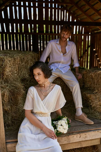 Mariée asiatique en robe blanche tenant bouquet et assis sur le foin près du marié roux dans la grange, rural — Photo de stock