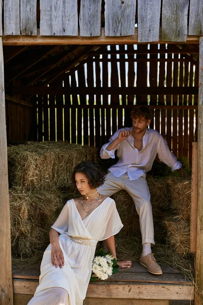Asian woman in wedding dress holding bouquet and sitting on hay near redhead groom in barn, rural — Stock Photo