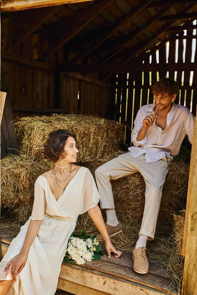 Happy asian bride in white dress holding bouquet and sitting on hay near groom in barn, rural — Stock Photo