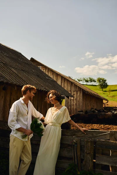 Happy asian bride in white dress standing near groom in sunglasses near stable in countryside — Stock Photo