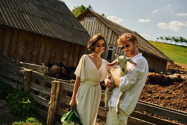 Lächelndes multiethnisches Paar in Brautkleid und Sonnenbrille, das niedliche Ziegenbaby kuschelt, Landschaft — Stockfoto