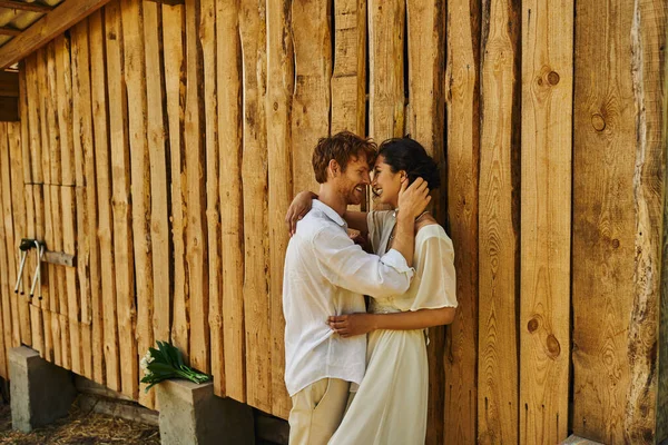 Boda estilo boho, novio feliz coqueteando con novia asiática en vestido blanco, recién casados en el campo - foto de stock