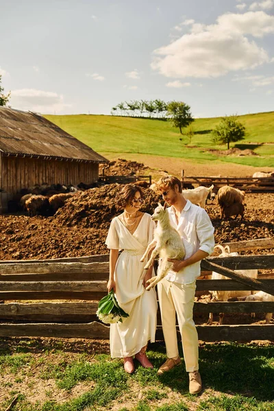 Mariage de style boho dans la campagne, marié heureux tenant bébé chèvre près de mariée asiatique en robe blanche — Photo de stock