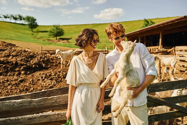 Just married couple, redhead groom holding baby goat near asian bride in white dress, boho style — Stock Photo