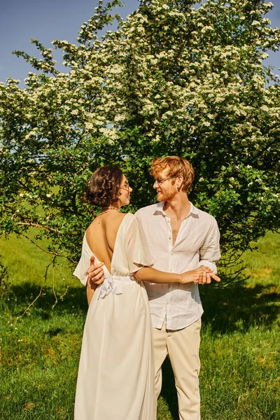 Feliz recién casados en el campo, novia asiática en vestido blanco y pelirroja novio abrazando cerca de gran árbol - foto de stock