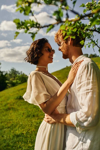 Boho-Stil, glückliches interrassisches Brautpaar in Sonnenbrille und Brautkleid, das sich im grünen Garten umarmt — Stockfoto