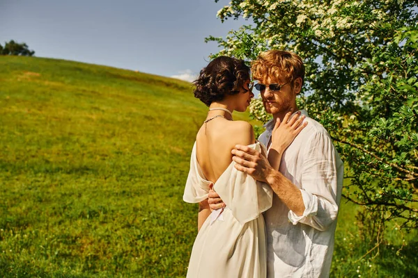 Estilo boho, feliz multiétnico recém-casados em óculos de sol e vestido de casamento abraçando no campo — Fotografia de Stock