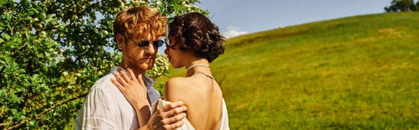 Estilo boho, feliz pareja multiétnica en gafas de sol y vestido de novia abrazándose en el campo, bandera - foto de stock