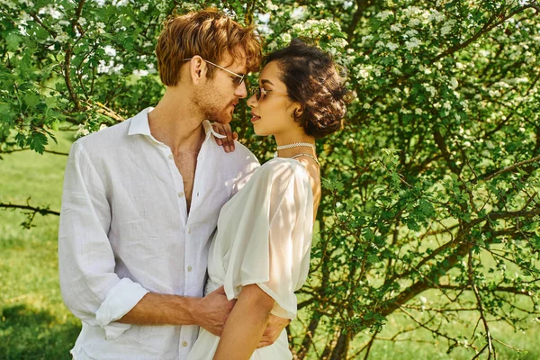 Feliz pareja multiétnica en gafas de sol y vestido de novia abrazándose en el campo, la vida de estilo boho - foto de stock